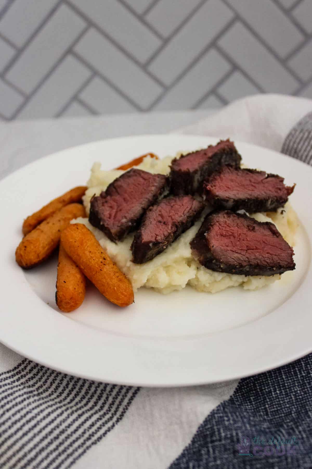 Boneless short ribs sliced over mashed potatoes with carrots on a white plate.