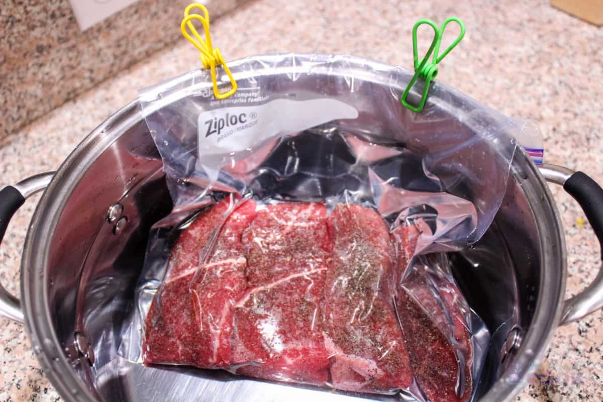 Sealed ziplock bag secured to the side of a large pot with chip clips, and the meat in the bag below the pot's water line.