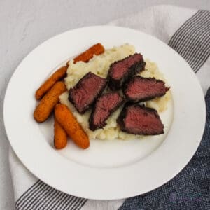 Boneless short ribs sliced over mashed potatoes with carrots on a white plate.