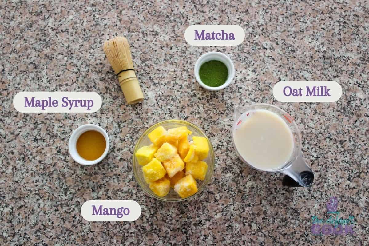 Labeled ingredients on a counter as follows: maple syrup, mango, matcha powder, and oat milk.