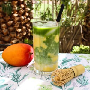 Mango, bamboo whisk, and clear glass of mango matcha latte on a towel outside near a palm tree.