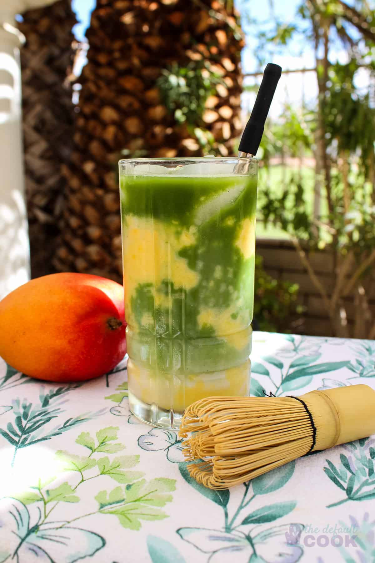 Mango, bamboo whisk, and clear glass of mango matcha latte on a towel outside near a palm tree.