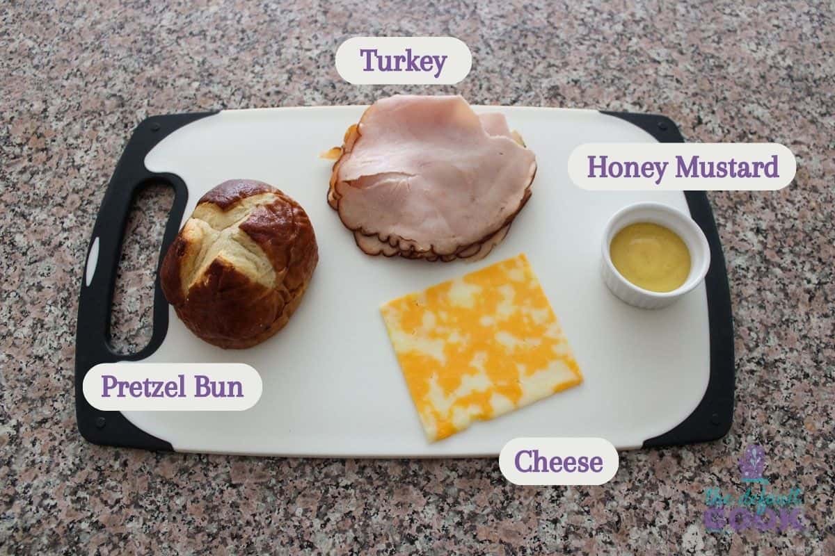 A cutting board on a counter with labeled ingredients resting on it: pretzel bun, colby cheese slice, sliced turkey, and honey mustard.
