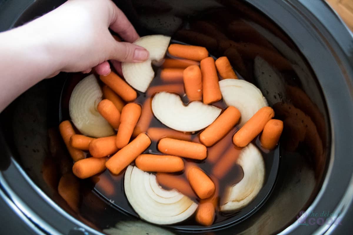 Carrots and onions in a crock pot with a hand placing the last onion section in.