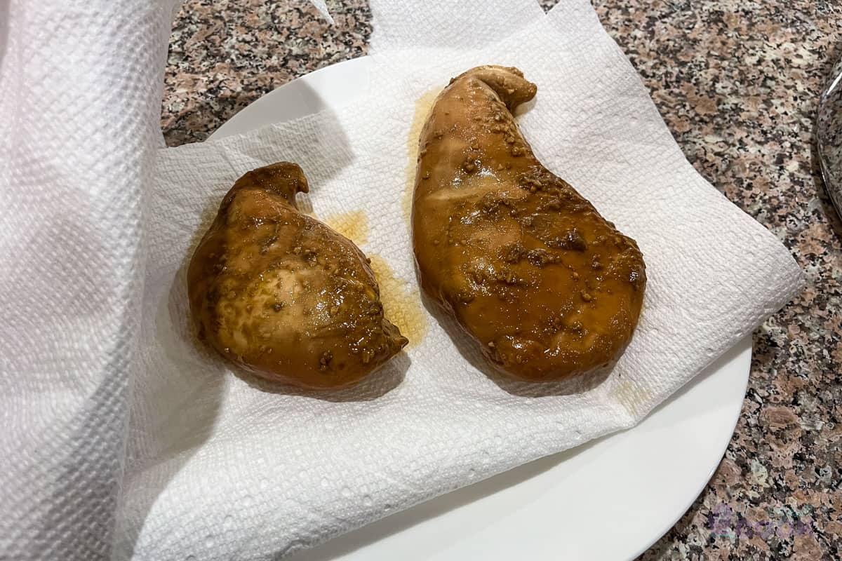 Two sous vide cooked chicken breasts on a paper towel on a plate, with another paper towel about to be laid on top.