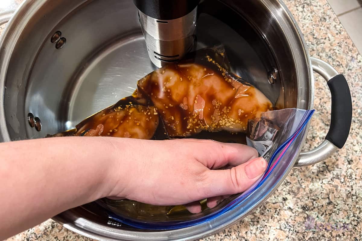 Chicken teriyaki in a zip top bag mostly submerged, with the zip top about two inches open and a hand pressing out the last of the air above the water line while closing it.