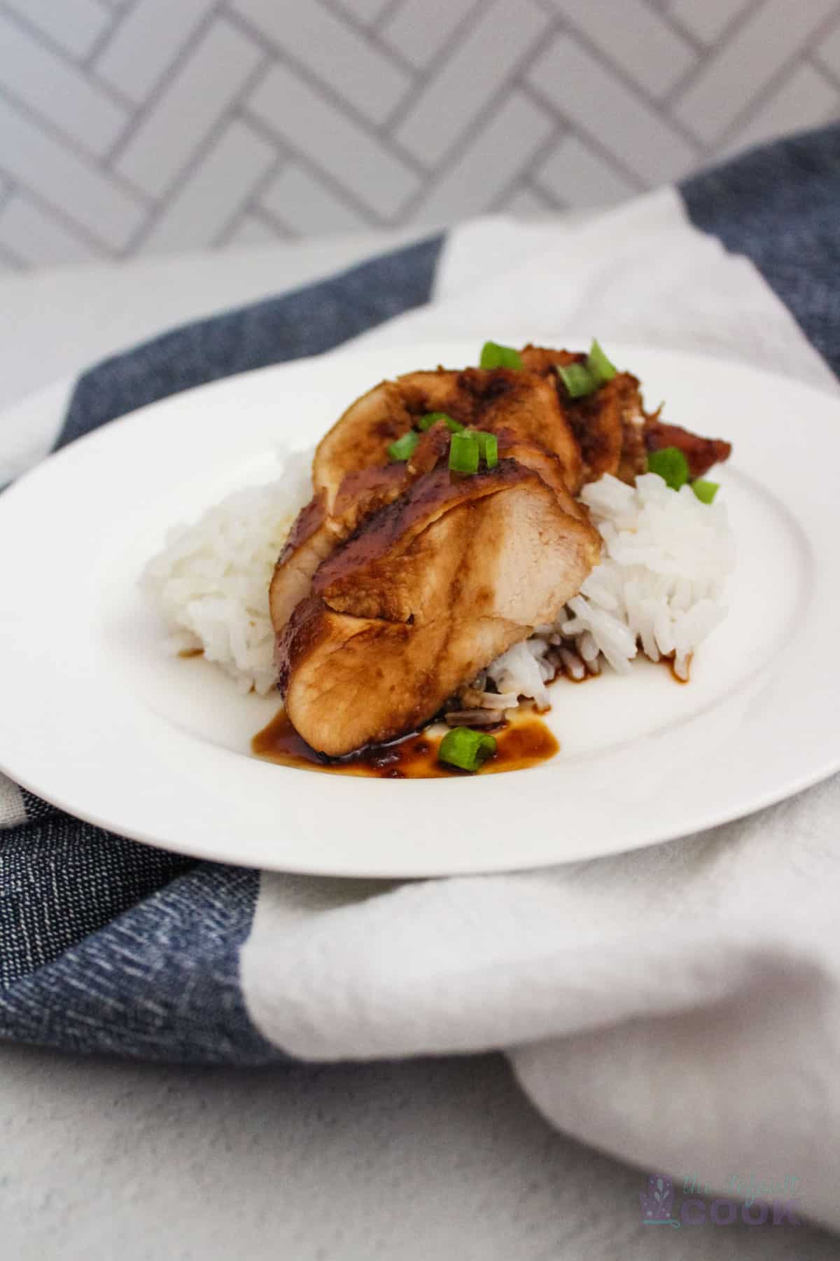 Plate of sliced chicken breast with teriyaki sauce over rice garnished with green onions on a kitchen towel.