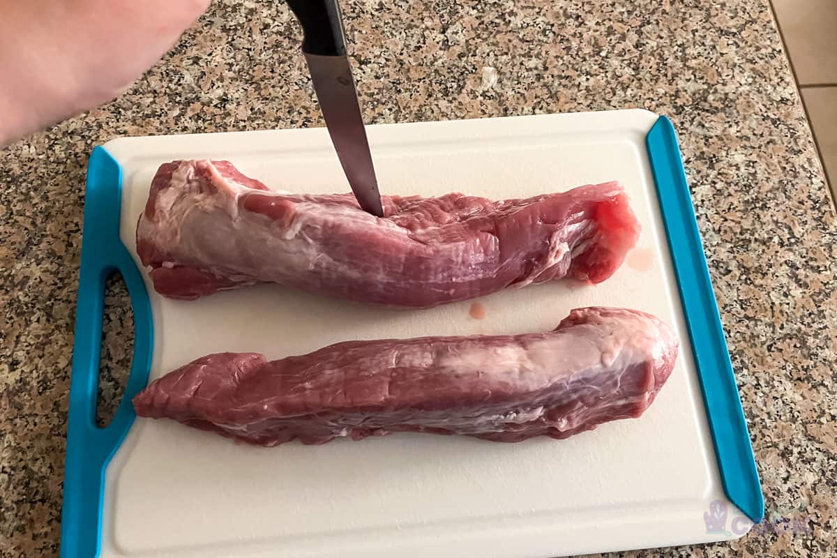 Piercing raw pork tenderloin with a knife on a cutting board.