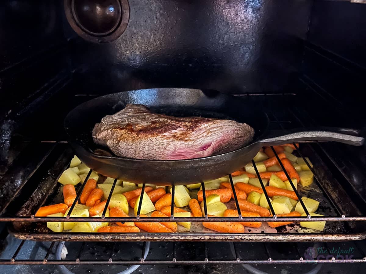 Cast iron skillet inside an oven on the top rack with a sheet pan of vegetables below it on the lower rack.