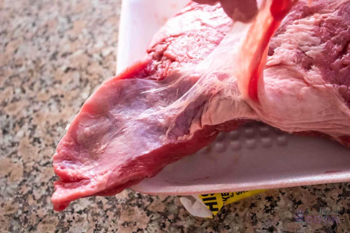 Close up of one end of a tri tip with a hand pulling the silver skin membrane away from the meat.