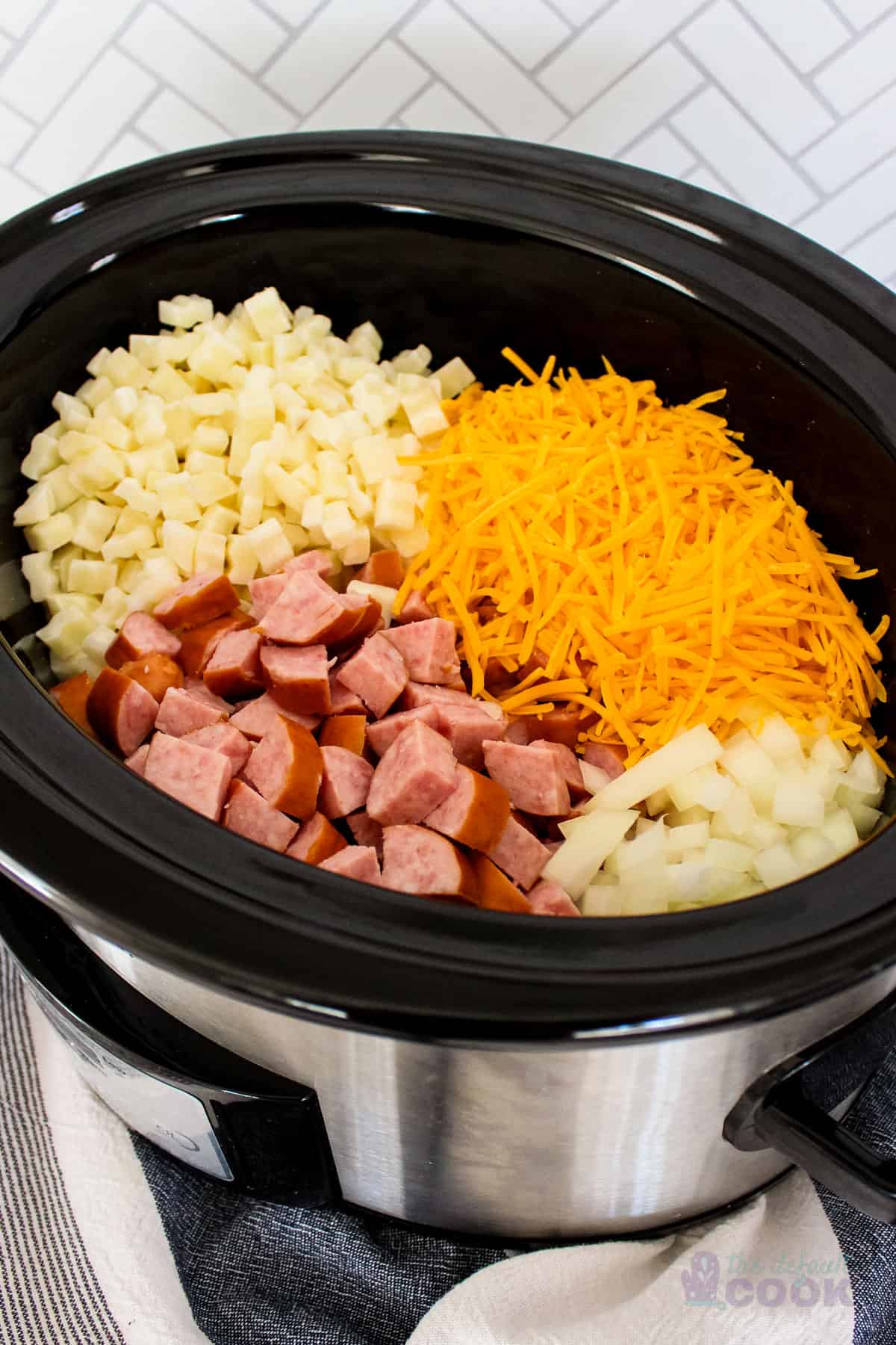 An oval crock pot with potatoes, kielbasa, onions, and cheddar cheese in it before cooking.