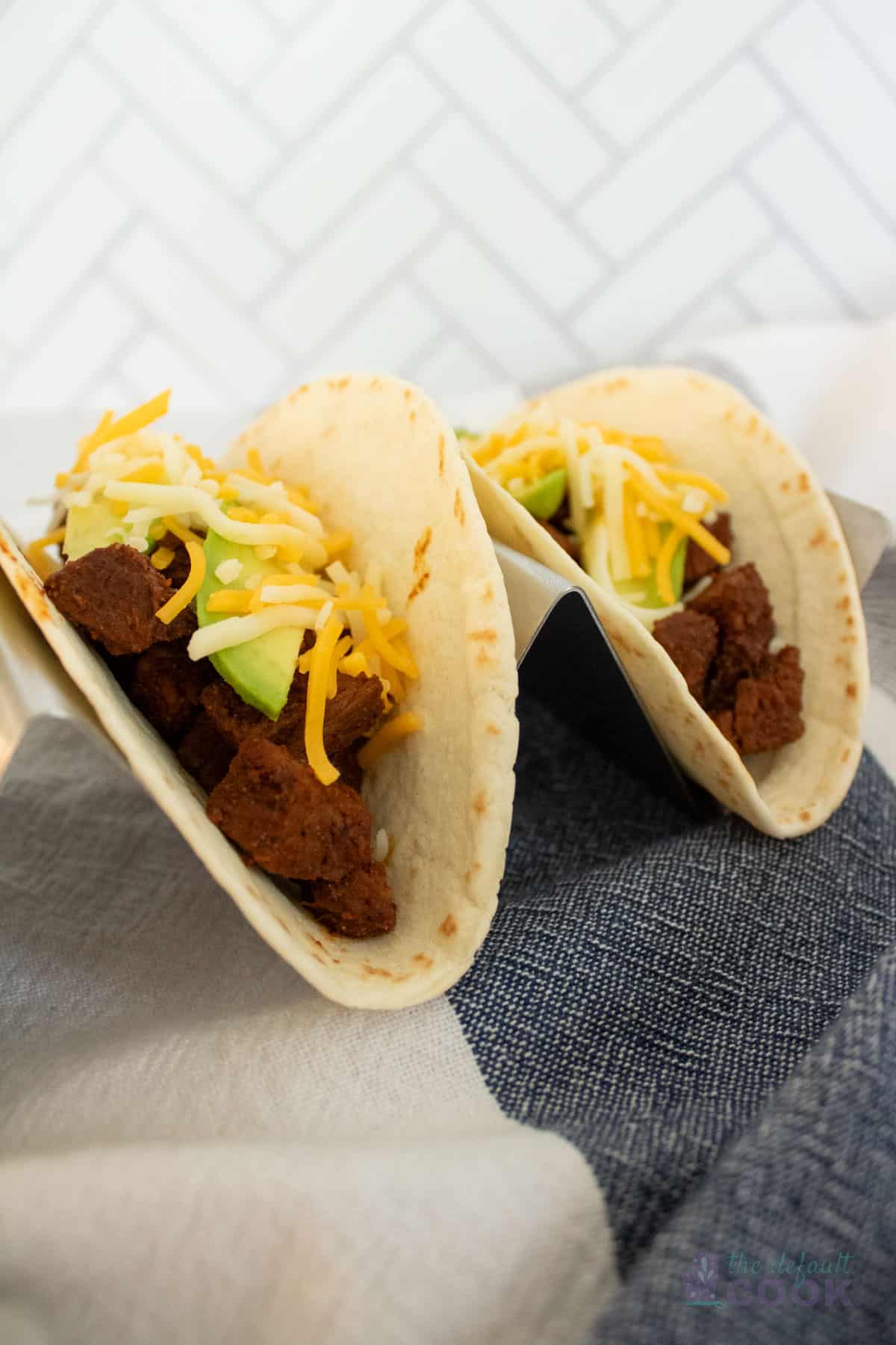 Two tri tip tacos topped with cheese and avocado in a taco holder on a blue and white kitchen towel.