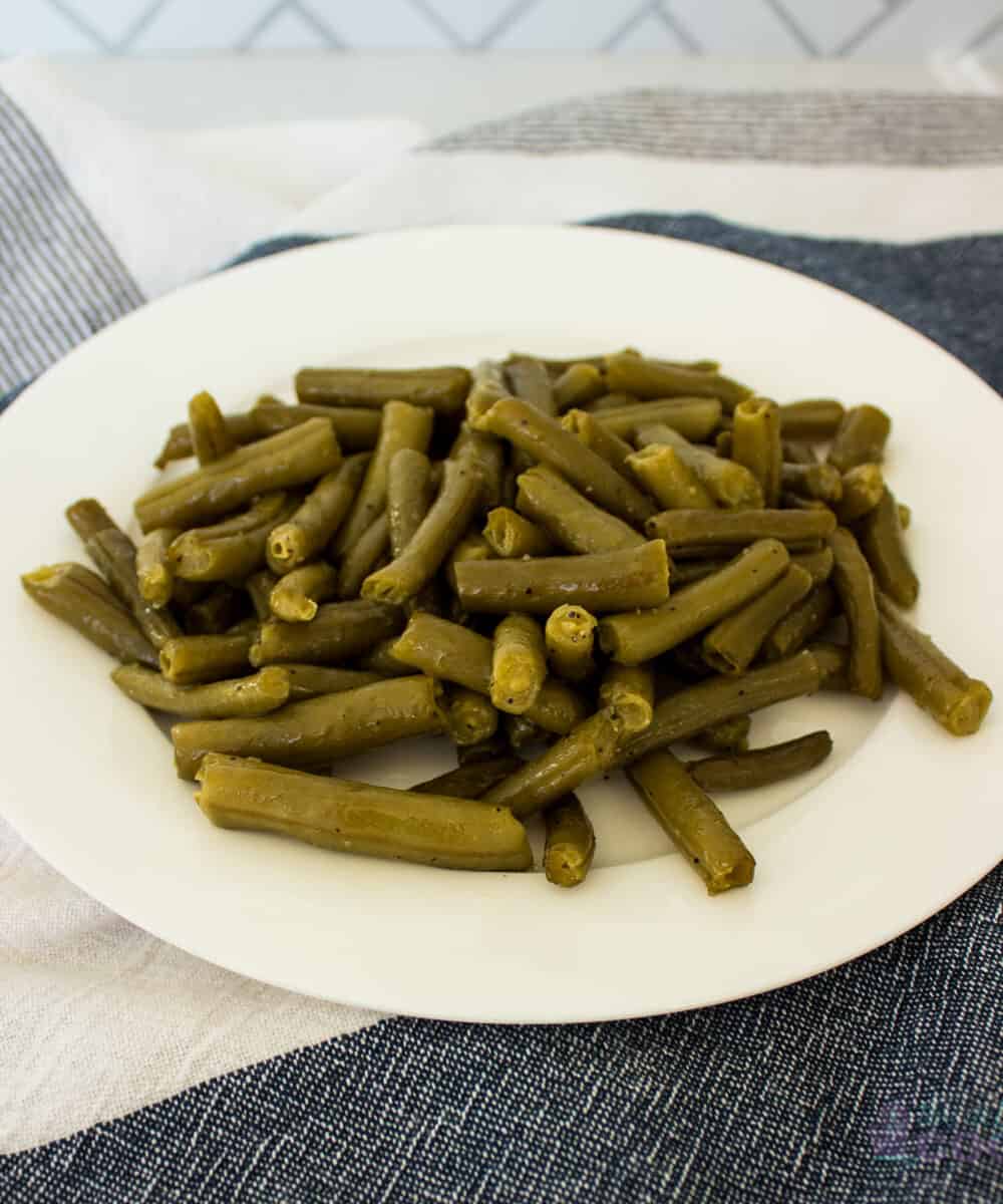 Cooked, seasoned green Beans on a white plate sitting on a blue and white kitchen towel.