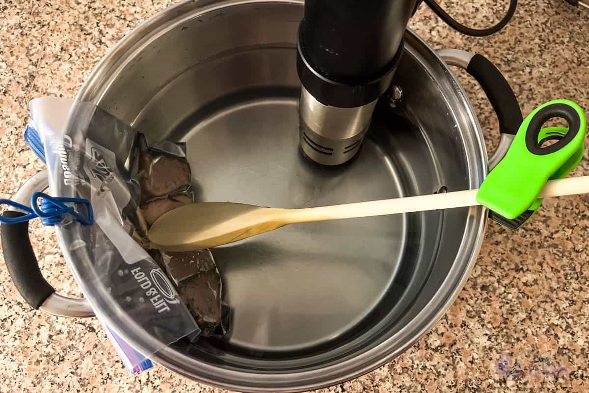 Pork slices in a sous vide bath to reheat, held in place by a chip clip and forced below the water line with a wooden spoon.
