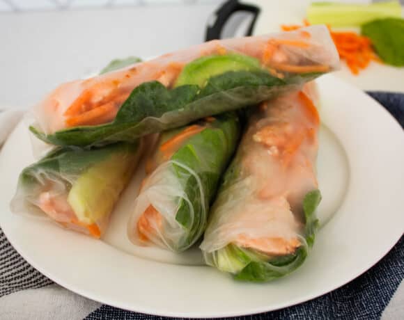 Four salmon spring rolls on a white plate with a kitchen towel under it and individual ingredients in the back.
