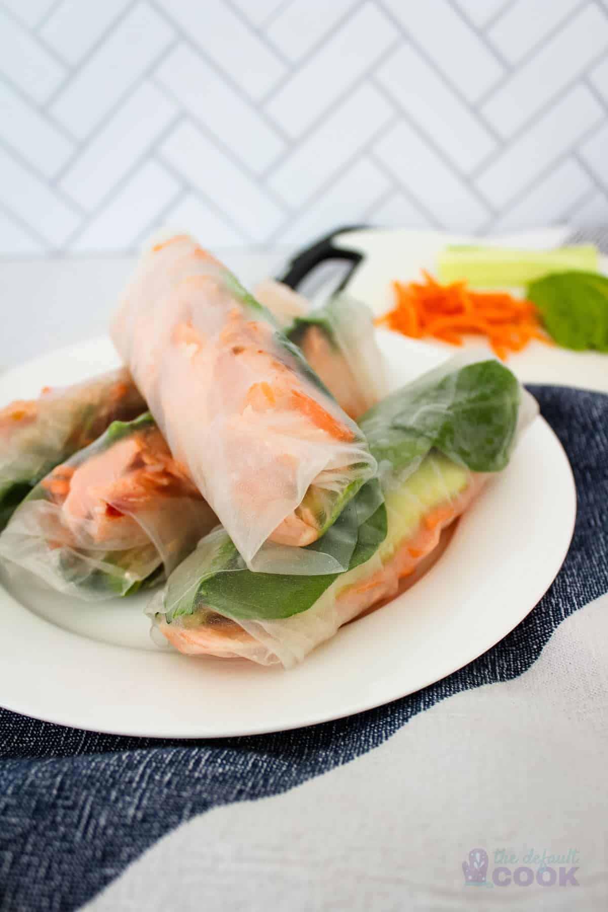 Four salmon spring rolls on a white plate with a kitchen towel under it and individual ingredients in the back.