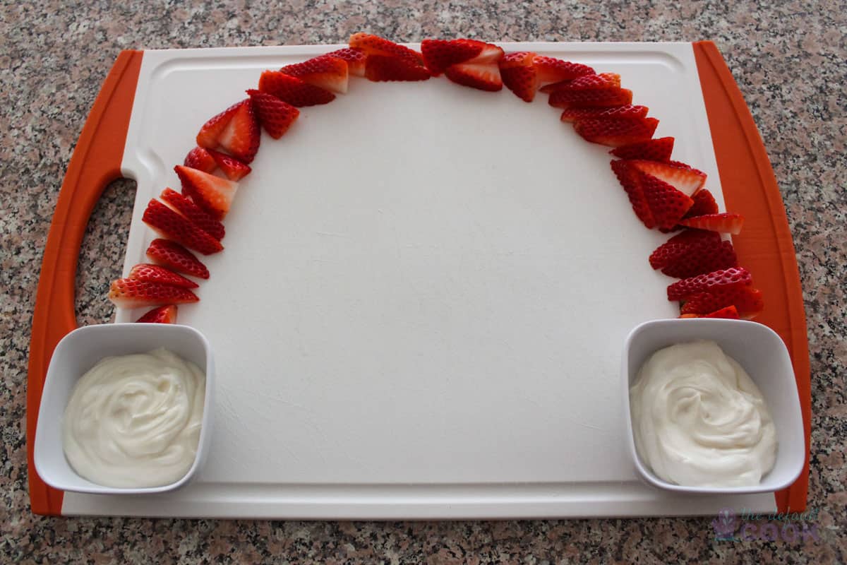 A cutting board on a counter with strawberries arching from one side up to the top and over to the other side, with yogurt dip in small bowls at the bottom of each side of the arch.