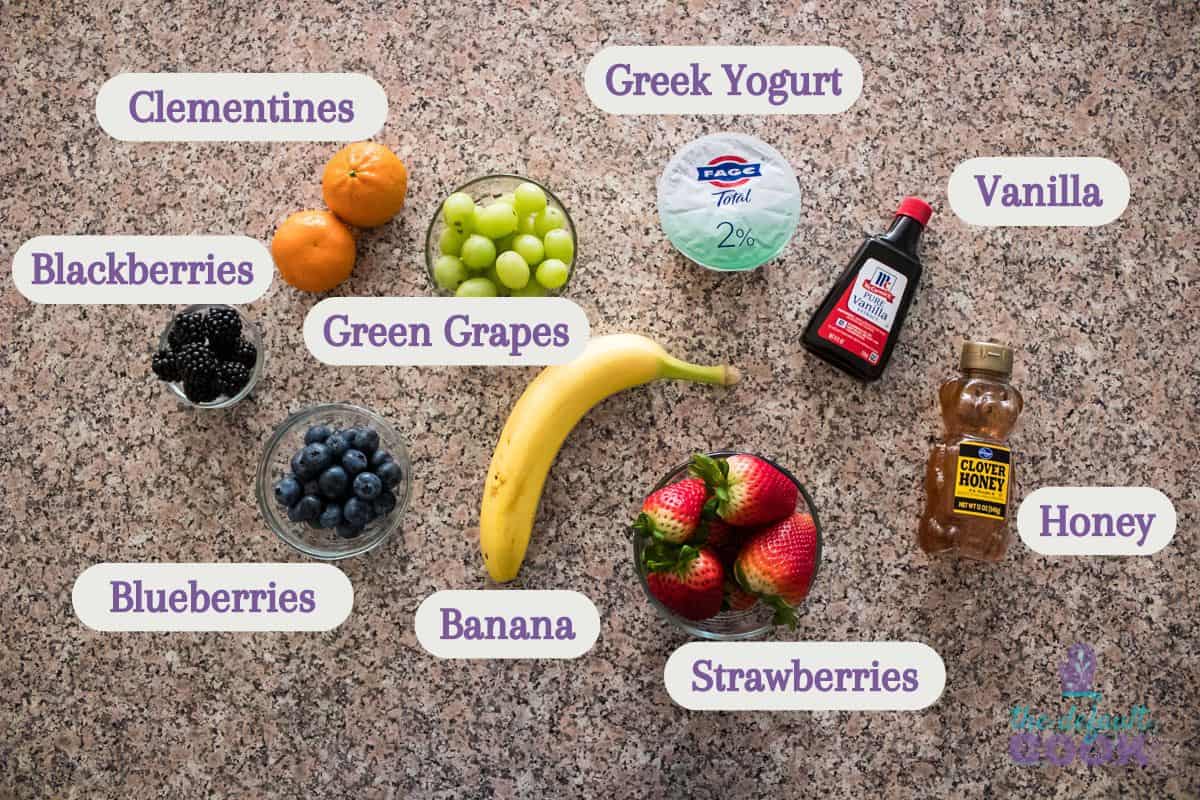 Assorted ingredients for a fruit platter spread out on a granite countertop, each labeled: Clementines, Blackberries, Green Grapes, Greek Yogurt, Vanilla, Honey, Blueberries, Banana, and Strawberries.