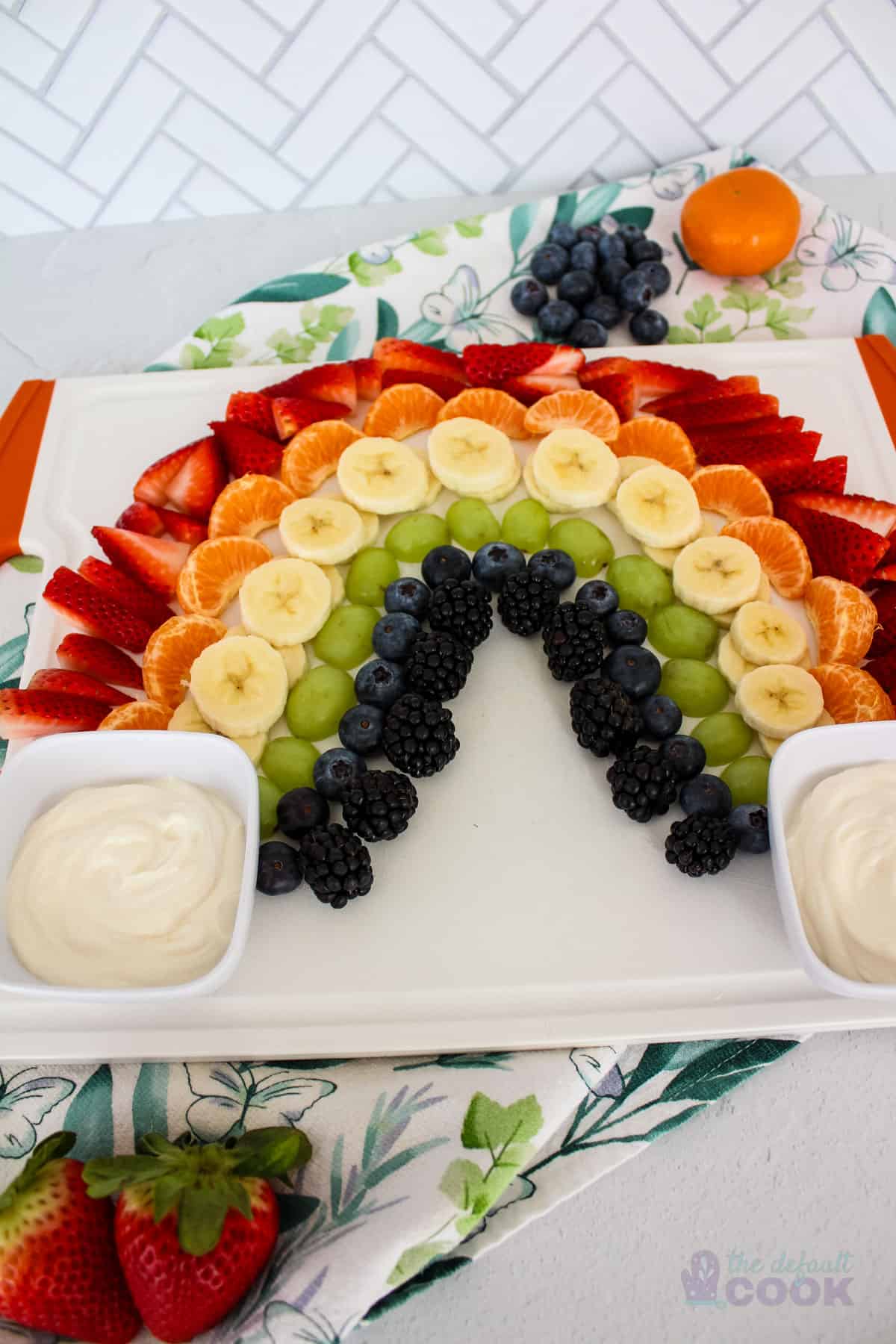 A fruit platter arranged in a rainbow pattern on a white serving tray, with yogurt dips on either side, against a white tile background with a decorative napkin, and 'the default cook' watermark at the bottom right.