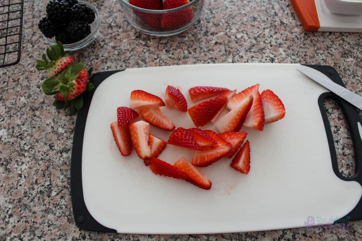 Sliced strawberries on a cutting board with other fruits and the display board partially in frame.