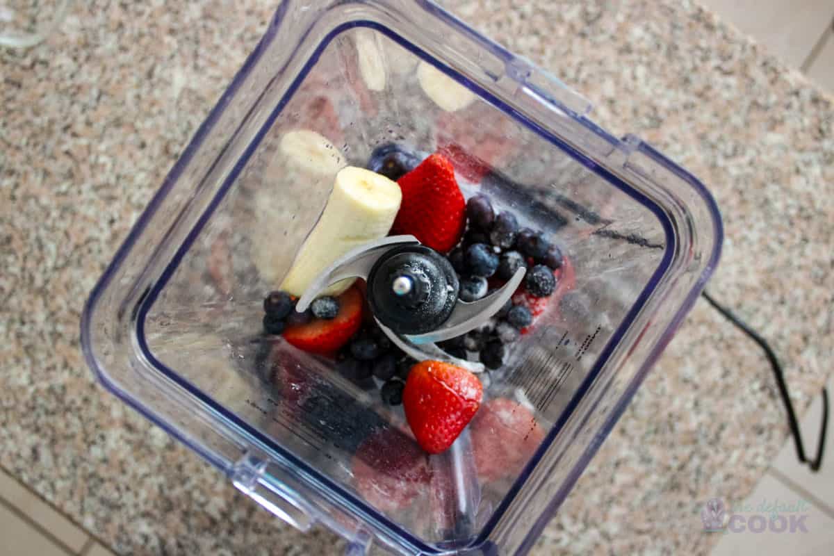 Top-down view into a blender containing fresh blueberries, sliced strawberries, and banana pieces, ready to be blended into a smoothie, with a kitchen countertop in the background.