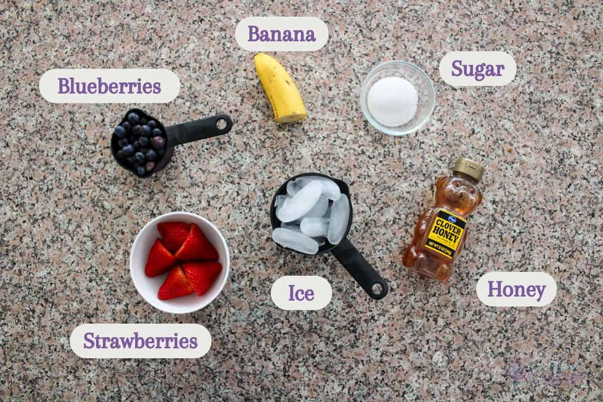 Smoothie ingredients laid out on a speckled kitchen countertop: a measuring cup of blueberries, a bowl of strawberries, half a banana, a measuring cup of ice, a small bowl of sugar, and a bottle of honey, each labeled with their respective names.