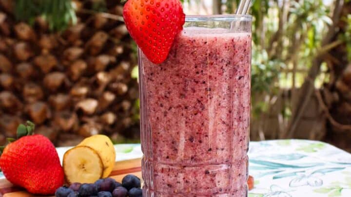 A glass filled with smoothie with a straw inserted and garnished with a fresh strawberry on the rim; in the background a small wooden cutting board with half a banana, blueberries, and a strawberry can be seen along with a lush tropical backdrop.