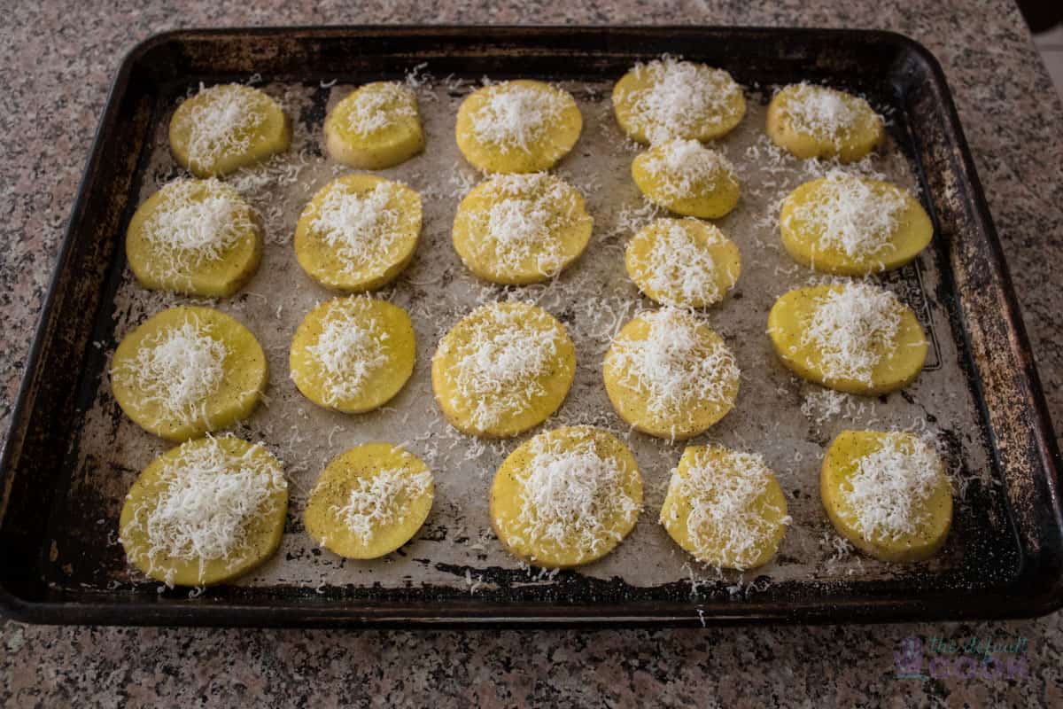 Seasoned potato slices seasoned and topped with parmesan cheese on a metal pan before going in the oven.