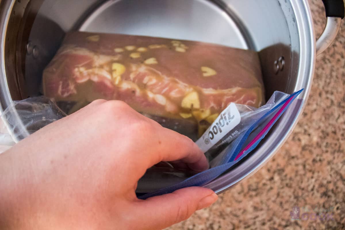 A hand is shown pressing a marinated pork loin inside a nearly sealed plastic bag to remove air before closing the bag and sous vide cooking the meat.