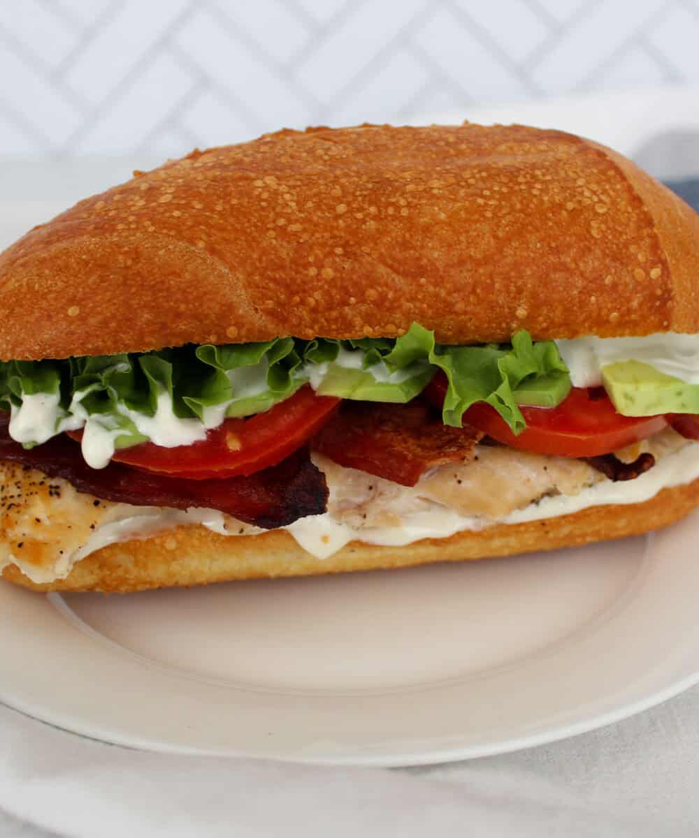 A chicken bacon ranch sandwich on a white plate on a kitchen towel with a tile background.