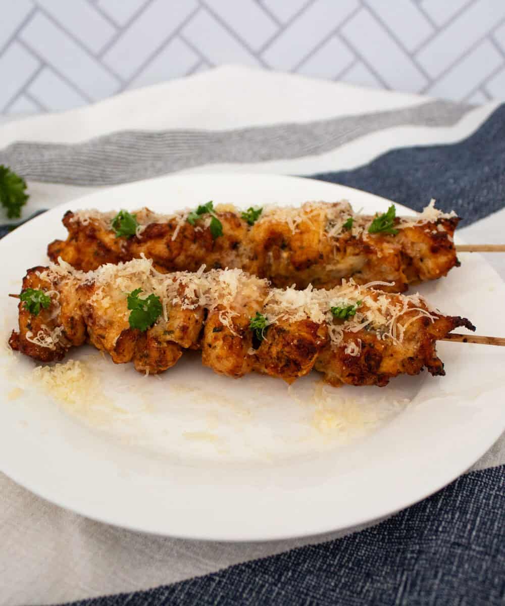 A plate with two chicken skewers on a kitchen towel, garnished with parmesan and parsley.