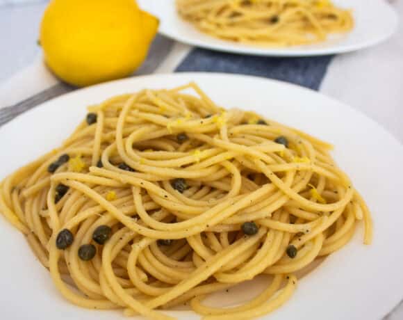 Two plates with spaghetti, capers, and lemon zest with a lemon between them and a garlic clove behind.