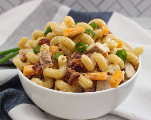 A bowl of ranch pasta salad made with cavatappi noodles, diced chicken breast and bacon, cheddar cheese cubes, and garnished with green onions. The bowl is set on blue and white striped cloth with a tiled background.