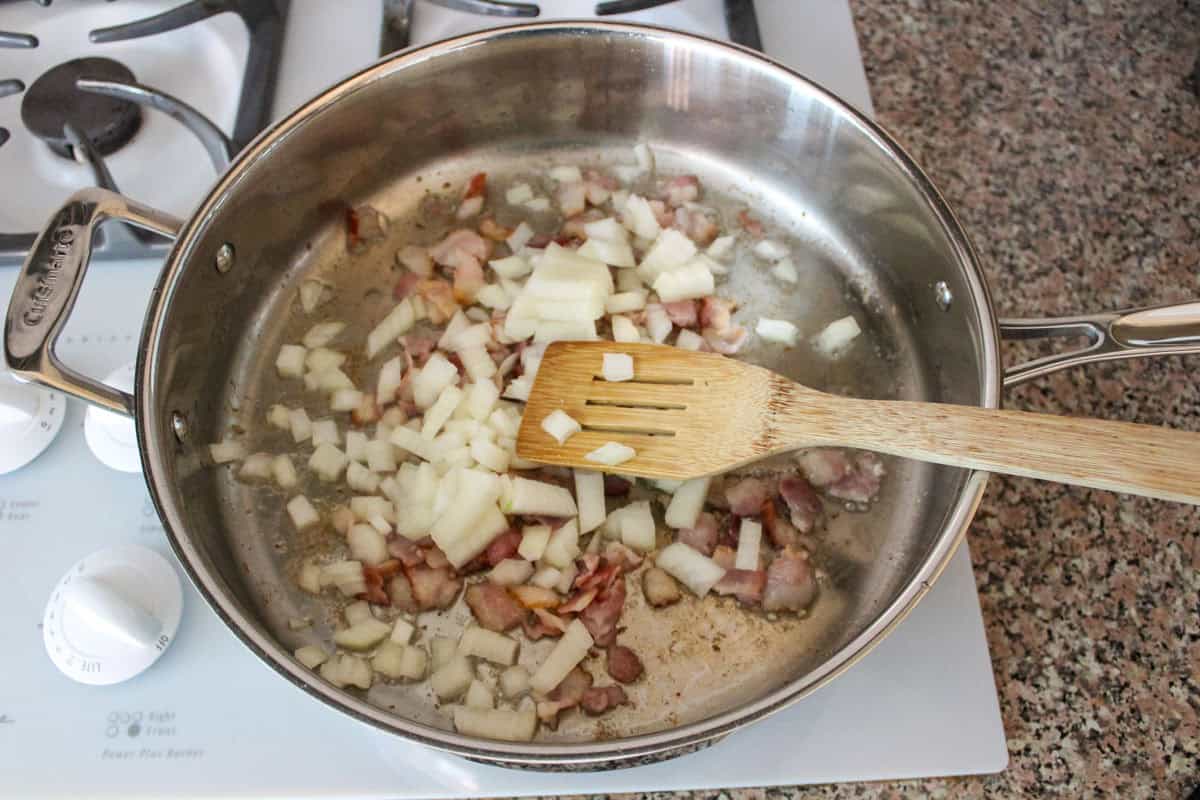 Chopped onions and pieces of bacon are being sautéed in a stainless steel pan on a stove, with a wooden spatula resting on top of the mixture.