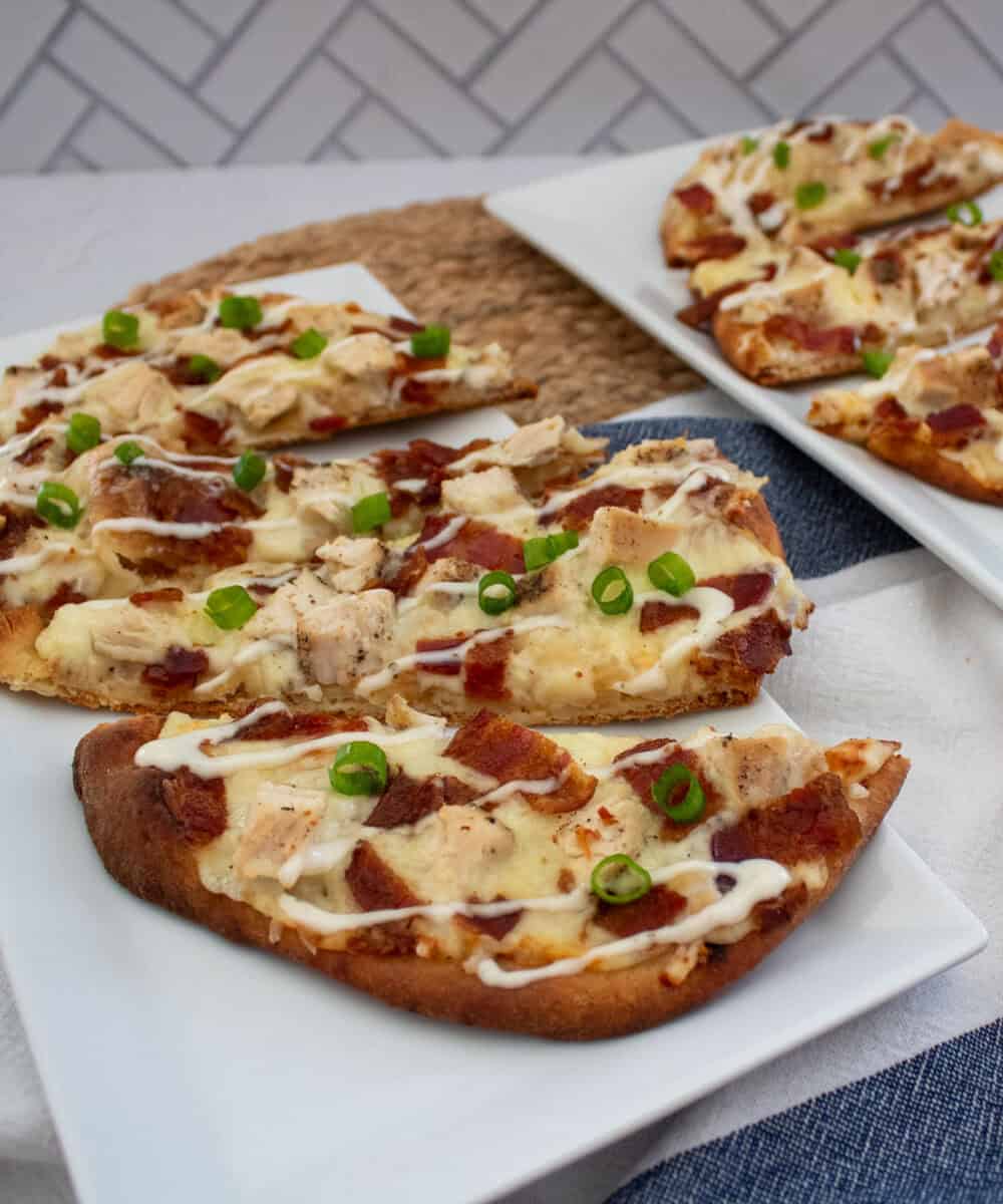 Flatbreads topped with chicken, bacon, melted cheese, and sliced green onions, drizzled with white sauce, arranged on rectangular white plates over a blue and white striped cloth.