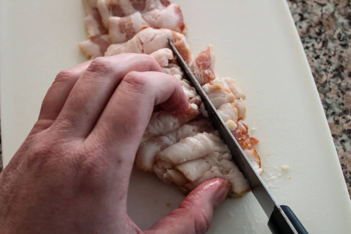 A hand is slicing raw bacon into pieces on a white cutting board with a kitchen knife.