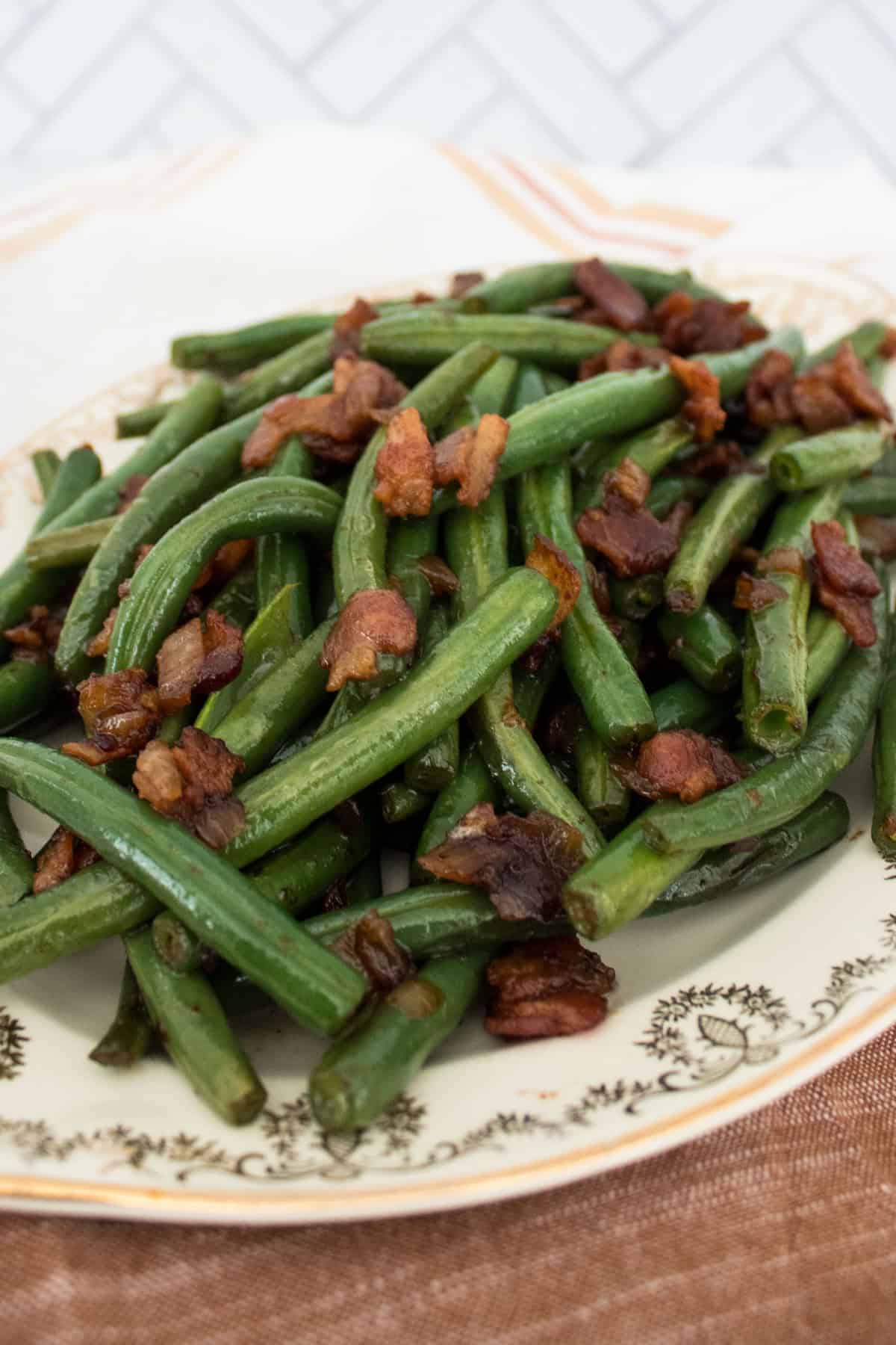 A vintage plate filled with cooked green beans mixed with crispy onion and bacon pieces, placed on a brown napkin with a white and brown towel in the background.