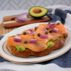 A slice of toast topped with smashed avocado, smoked salmon, red onion pieces, and capers on a plate. In the background are a halved avocado and sliced red onion on a wooden board.
