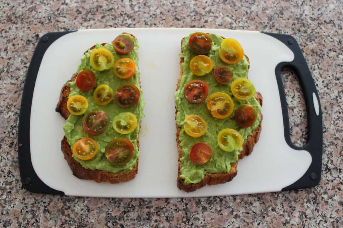 Two slices of toasted bread topped with mashed avocado and halved cherry tomatoes in red, yellow, and green, placed on a white cutting board with a black handle on a speckled countertop.