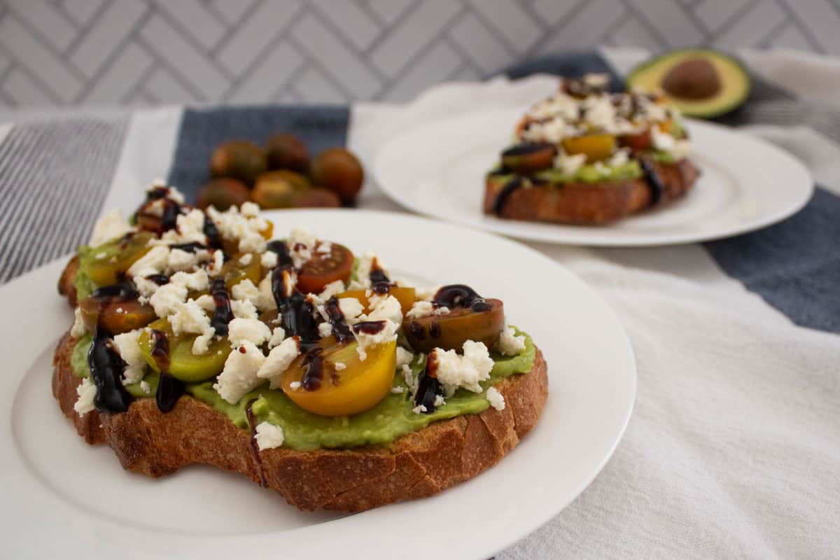 Sliced bread topped with smashed avocado, halved tomatoes, feta cheese, and balsamic glaze on a white plate, with more toast and an avocado in the background.