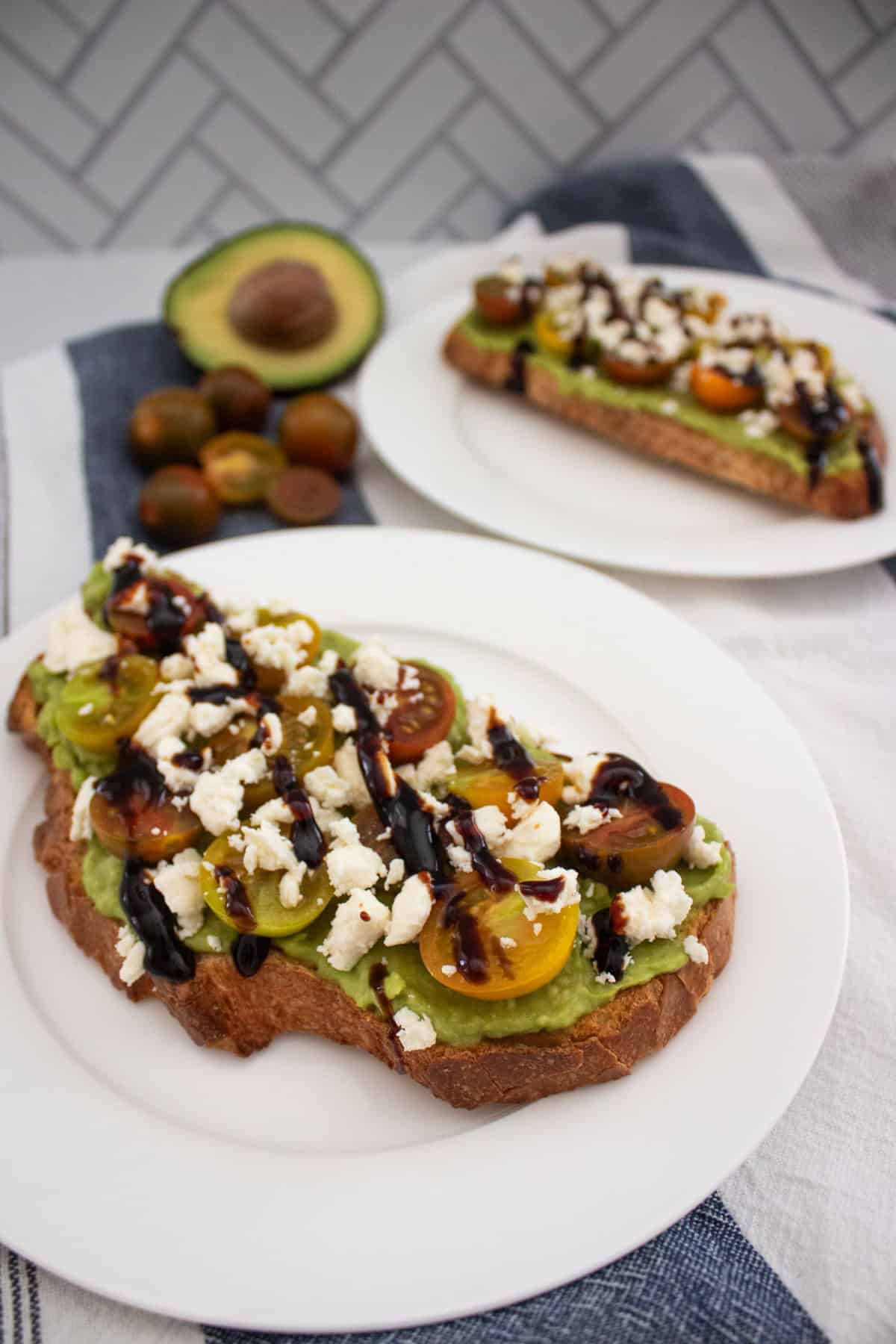 A slice of toast topped with mashed avocado, halved cherry tomatoes, crumbled cheese, and a drizzle of balsamic glaze on a white plate. Another similar toast, avocado, and tomatoes are in the background.