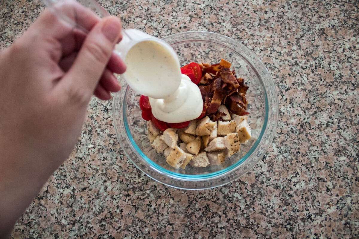 A hand pours creamy dressing from a measuring cup into a glass bowl filled with chopped grilled chicken, cherry tomatoes, and crumbled bacon on a speckled countertop.