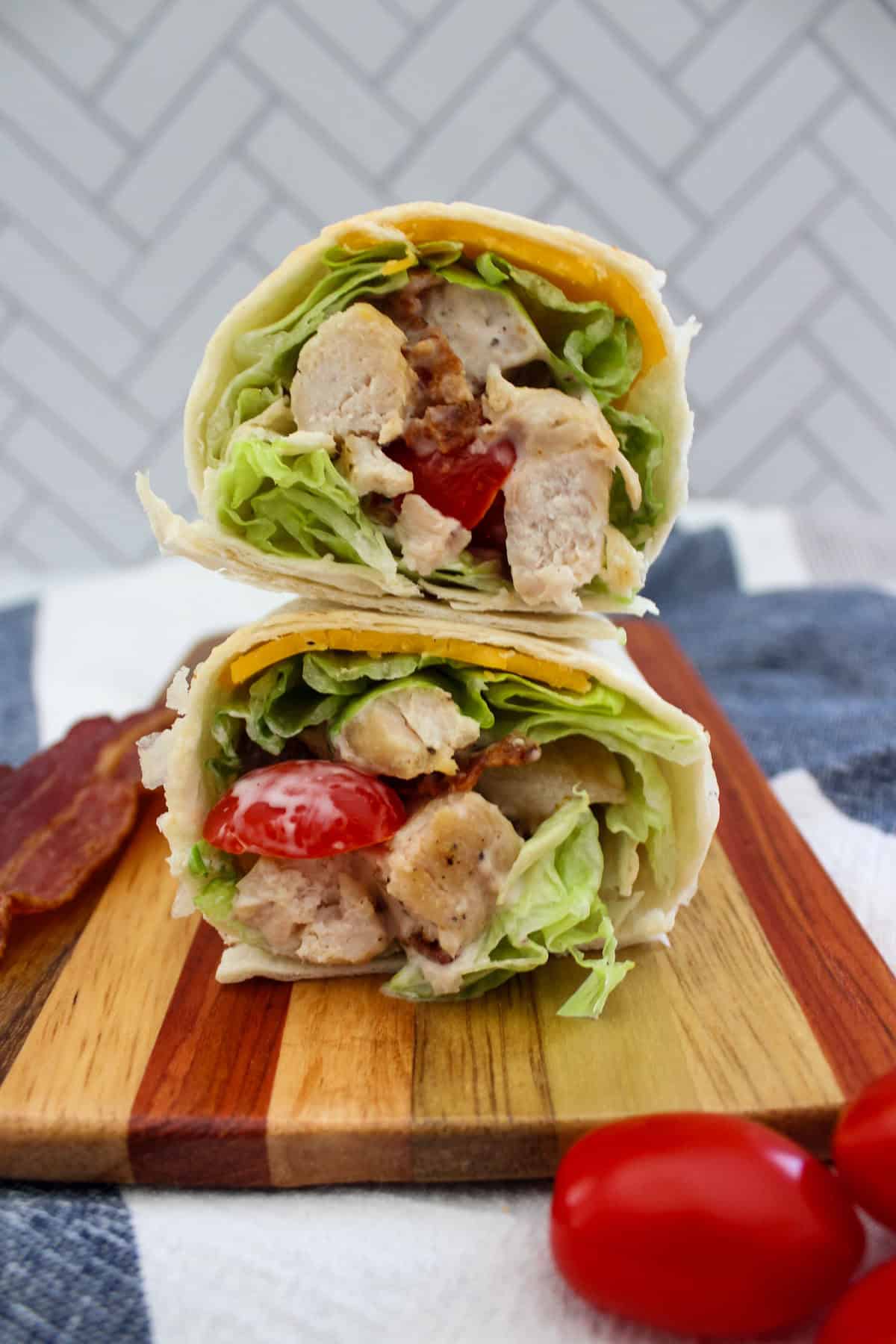 A wrap cut in half, filled with grilled chicken, lettuce, tomato, cheese, and bacon, stacked on a wooden board with cherry tomatoes and a bacon strip nearby. The background shows a white tiled wall.