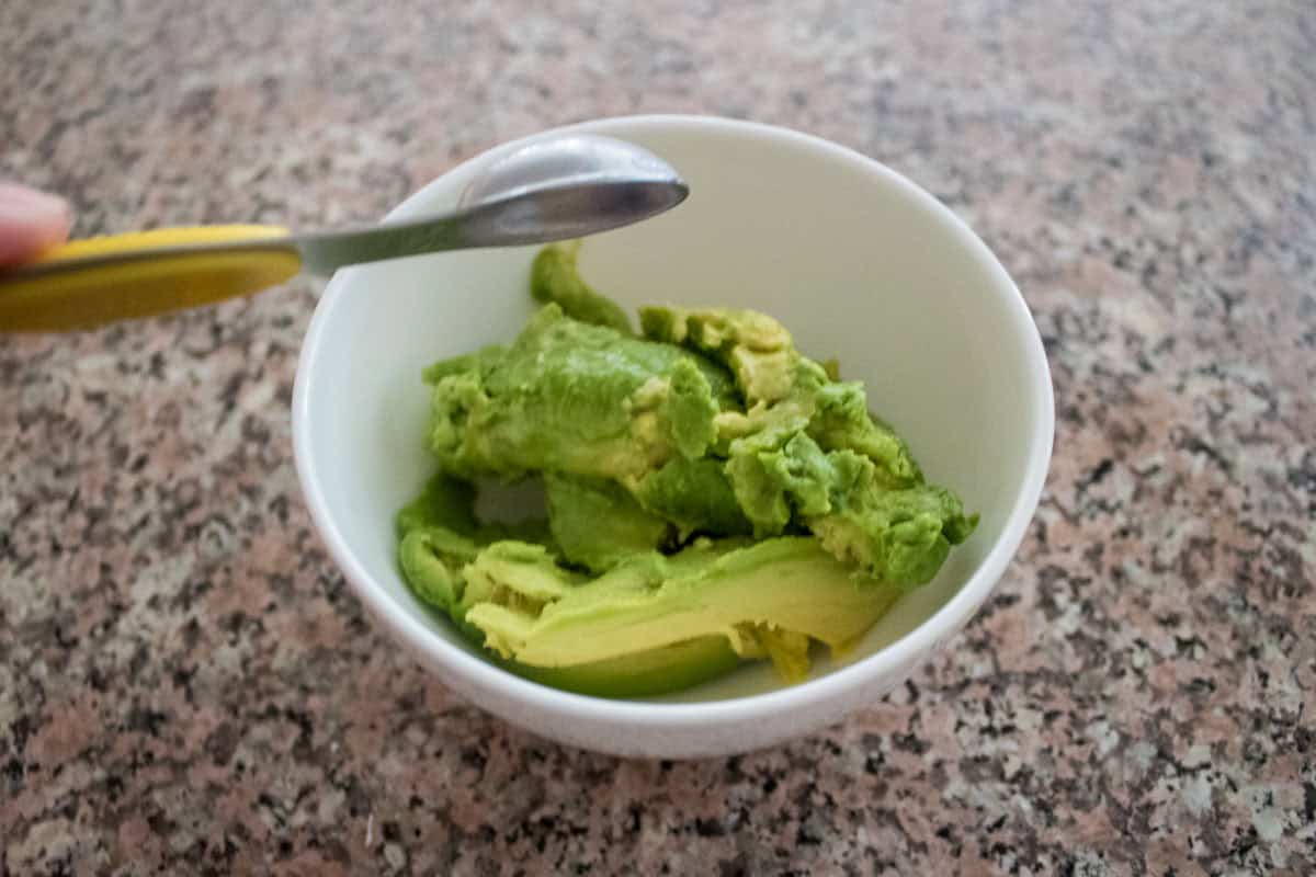A bowl containing mashed avocado sits on a granite countertop, with a spoon resting on the edge of the bowl.