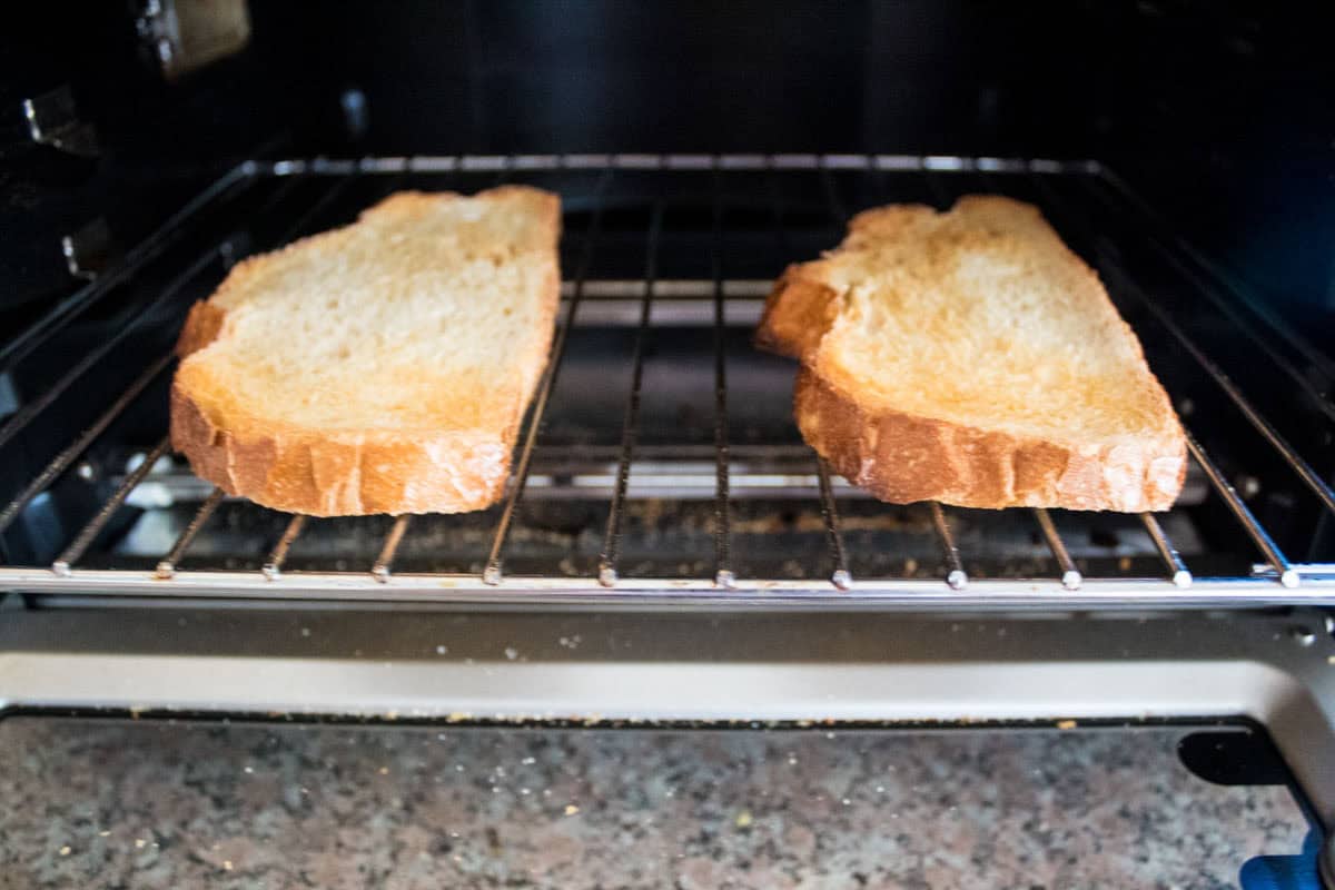 Two slices of bread being toasted directly on the wire rack inside an oven, with a granite countertop visible below the open oven door.