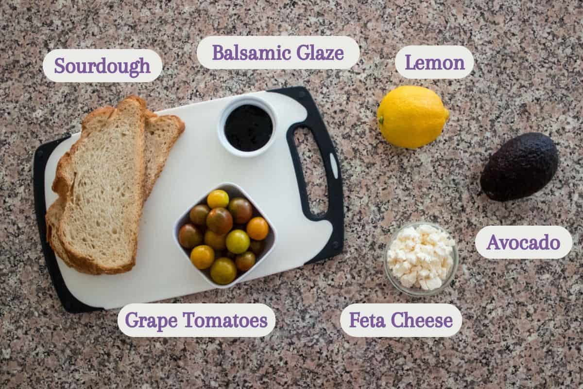Top-down view of ingredients on a countertop: slices of sourdough, a bowl of grape tomatoes, balsamic glaze, a lemon, an avocado, and a bowl of feta cheese, each labeled with text.