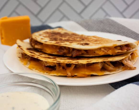 Two quesadillas filled with melted cheese, bacon, and shredded chicken are stacked on a white plate. A small bowl of ranch sauce sits in the foreground, with a block of cheddar cheese in the background.