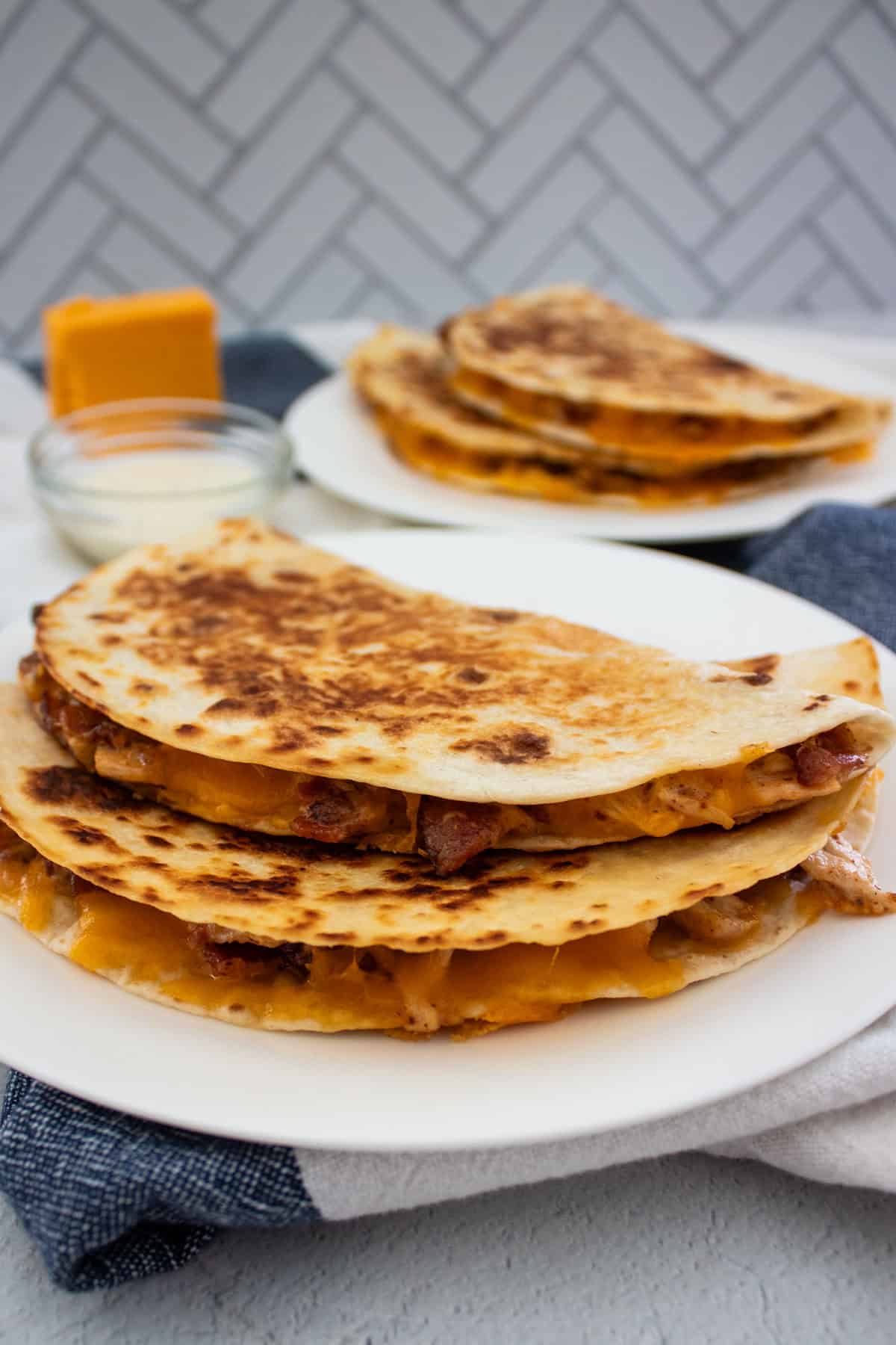 Two golden-brown quesadillas filled with melted cheese, chicken, and bacon on a white plate, with more quesadillas and a block of cheddar cheese in the background.