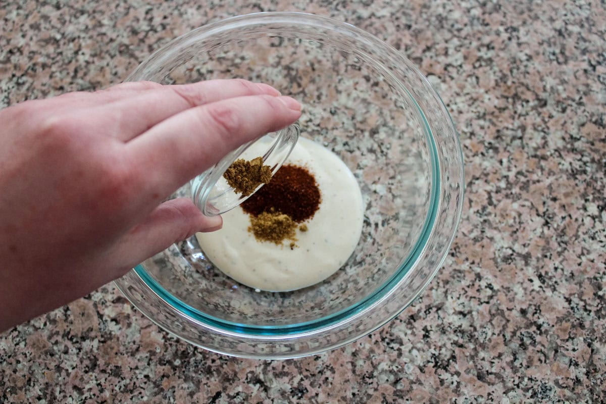 A hand pours ground spices from a small glass bowl into a larger glass bowl containing ranch dressing, set on a speckled countertop.