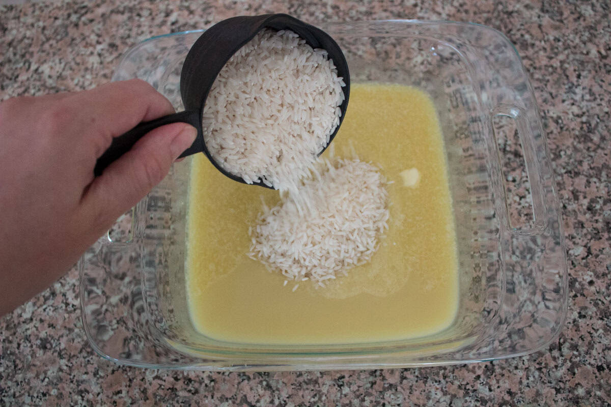 A hand pours a cup of uncooked white rice into a glass baking dish containing a yellow liquid on a speckled countertop.